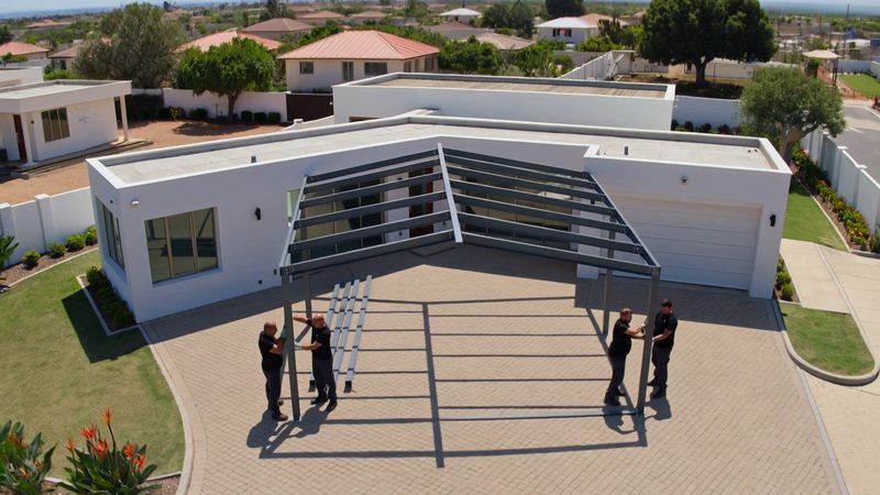 Four installers in black work clothes bolting the roof beams of a steel carport frame over a paved driveway
